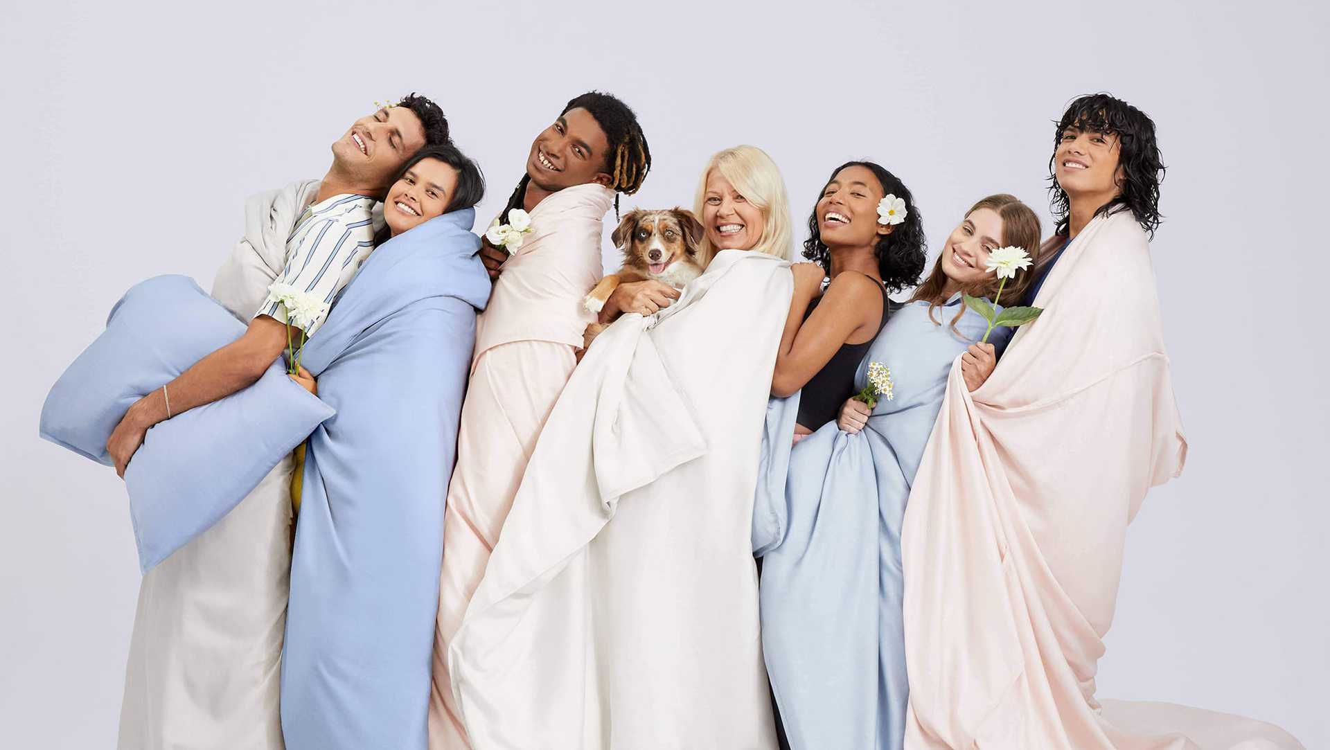 Diverse group of people holding flowers and a dog, each wrapped in sheets and comforters.